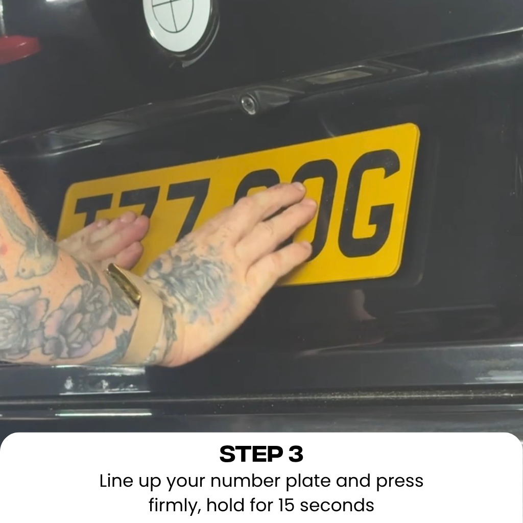 A close-up of a person's tattooed hand pressing down on a yellow UK license plate mounted on a black car, with instructional text at the bottom reading 'STEP 3 Line up your number plate and press firmly, hold for 15 seconds'.