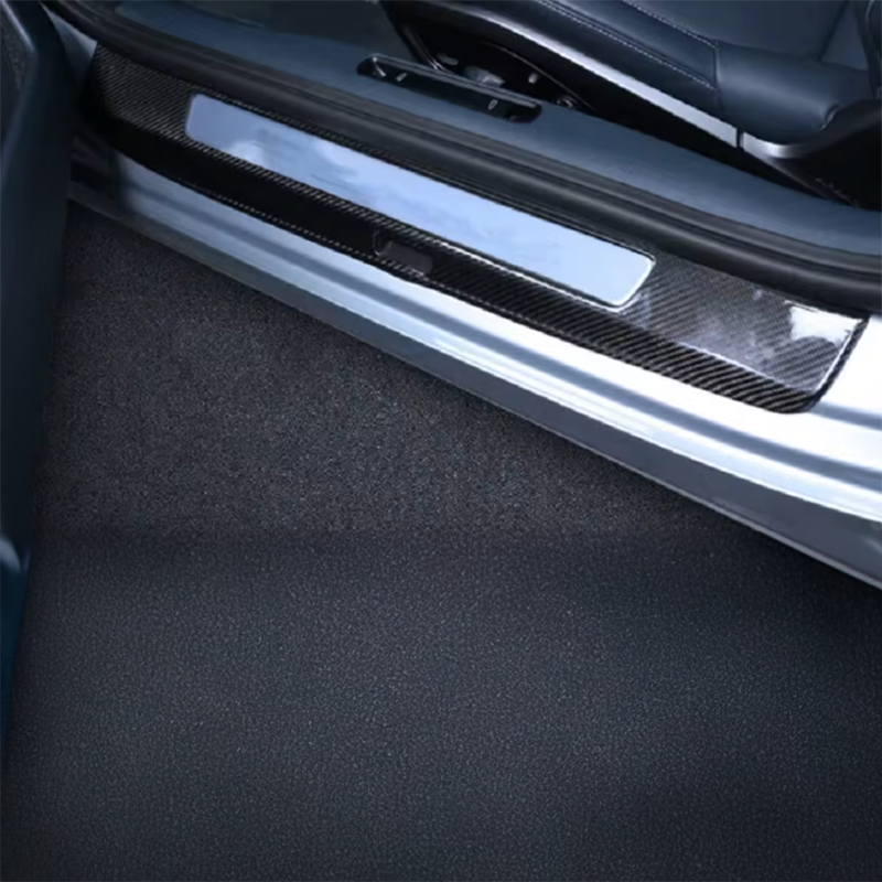A close-up of a car door sill trim featuring a carbon fiber pattern insert and a brushed silver metal accent, installed on a silver vehicle door frame with dark carpeted flooring.
